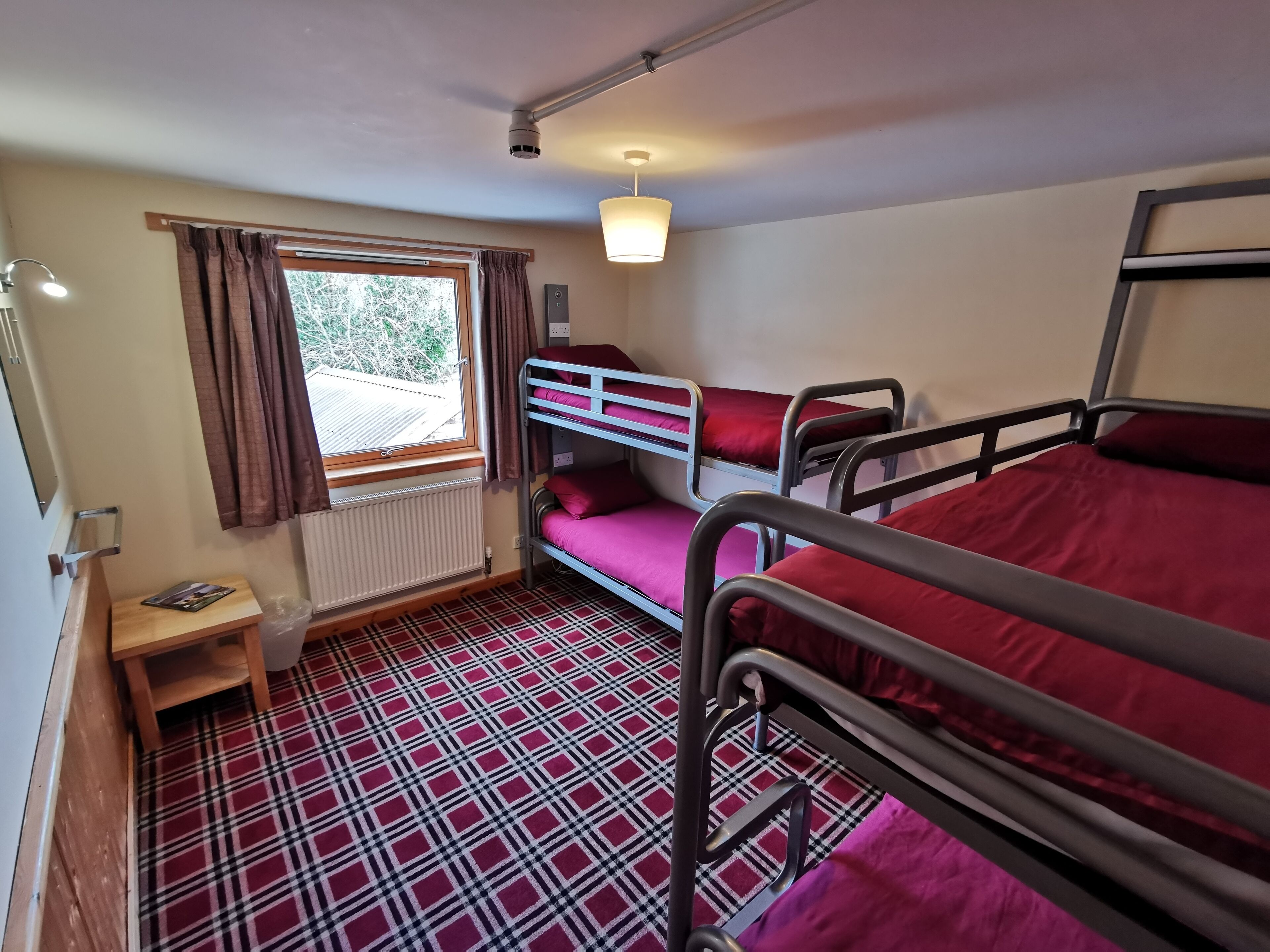 A dormitory room with bunk beds, a window with curtains, a radiator, a small wooden table, and a checkered floor.