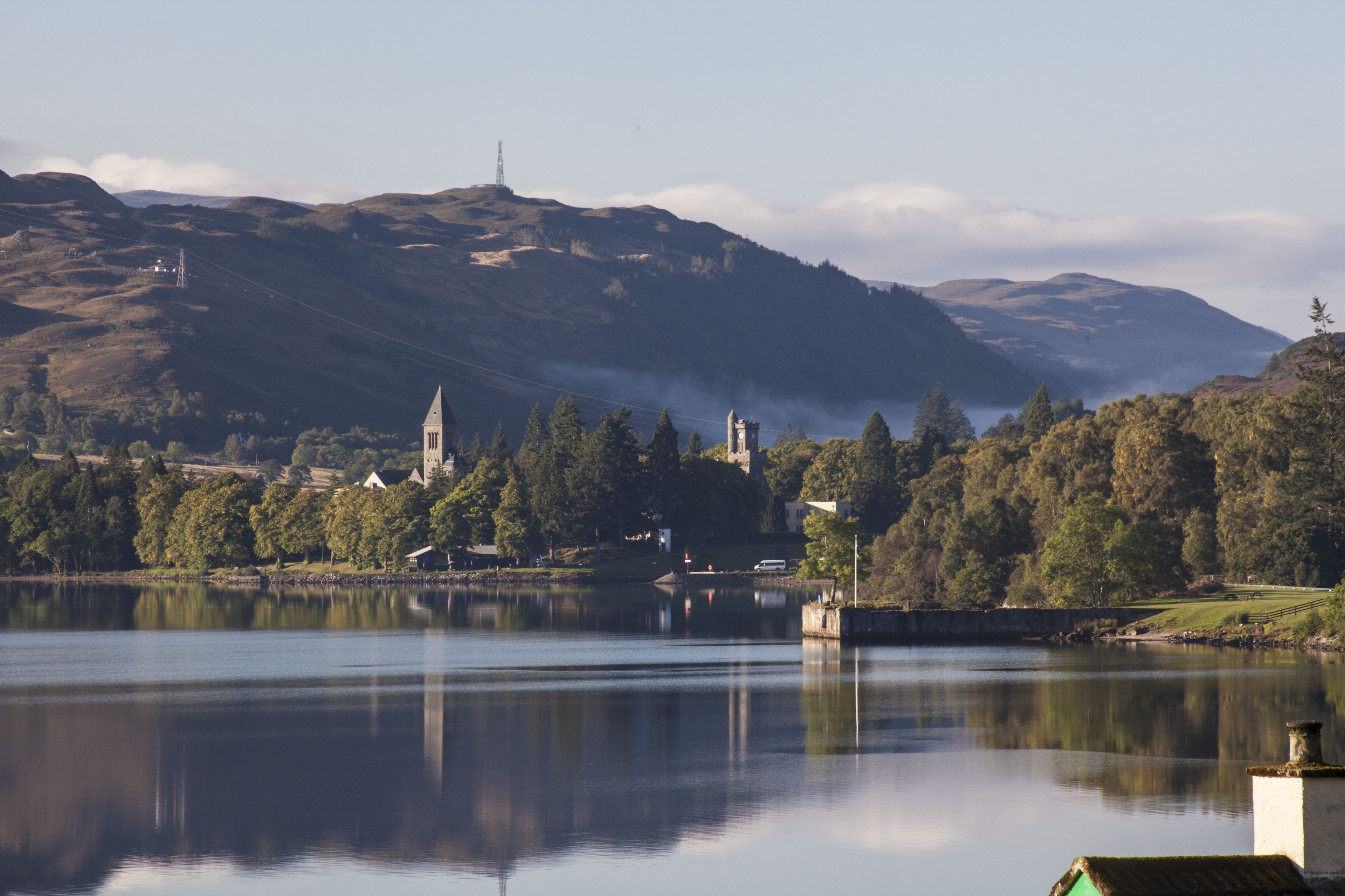 A calm lake with a church and a tower on a hill, surrounded by trees and hills.
