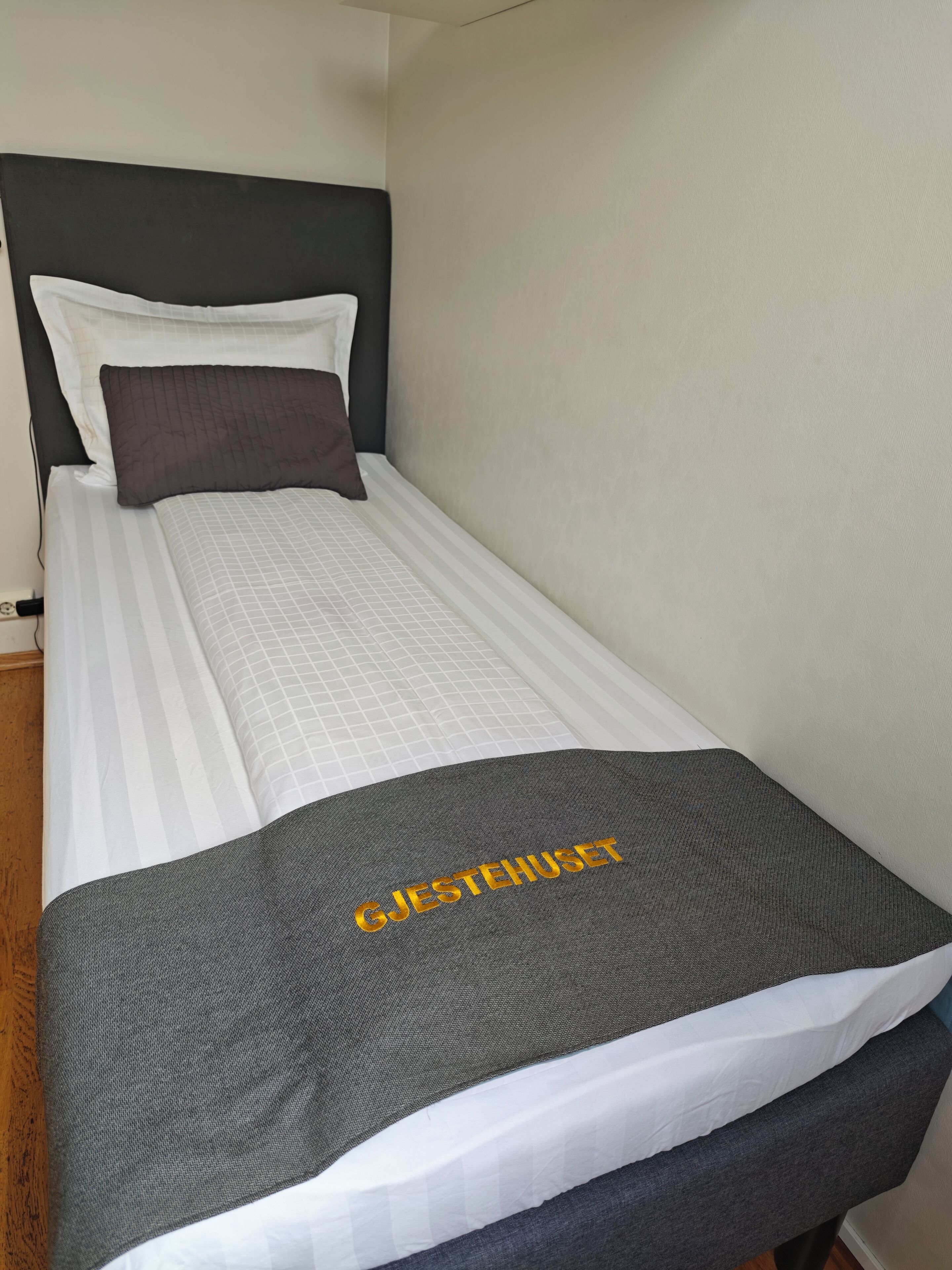A single bed with a dark headboard, white and gray bedding, and a gray blanket with 'GJESTEHUSET' text.