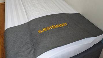 A single bed with a dark headboard, white and gray bedding, and a gray blanket with 'GJESTEHUSET' text.