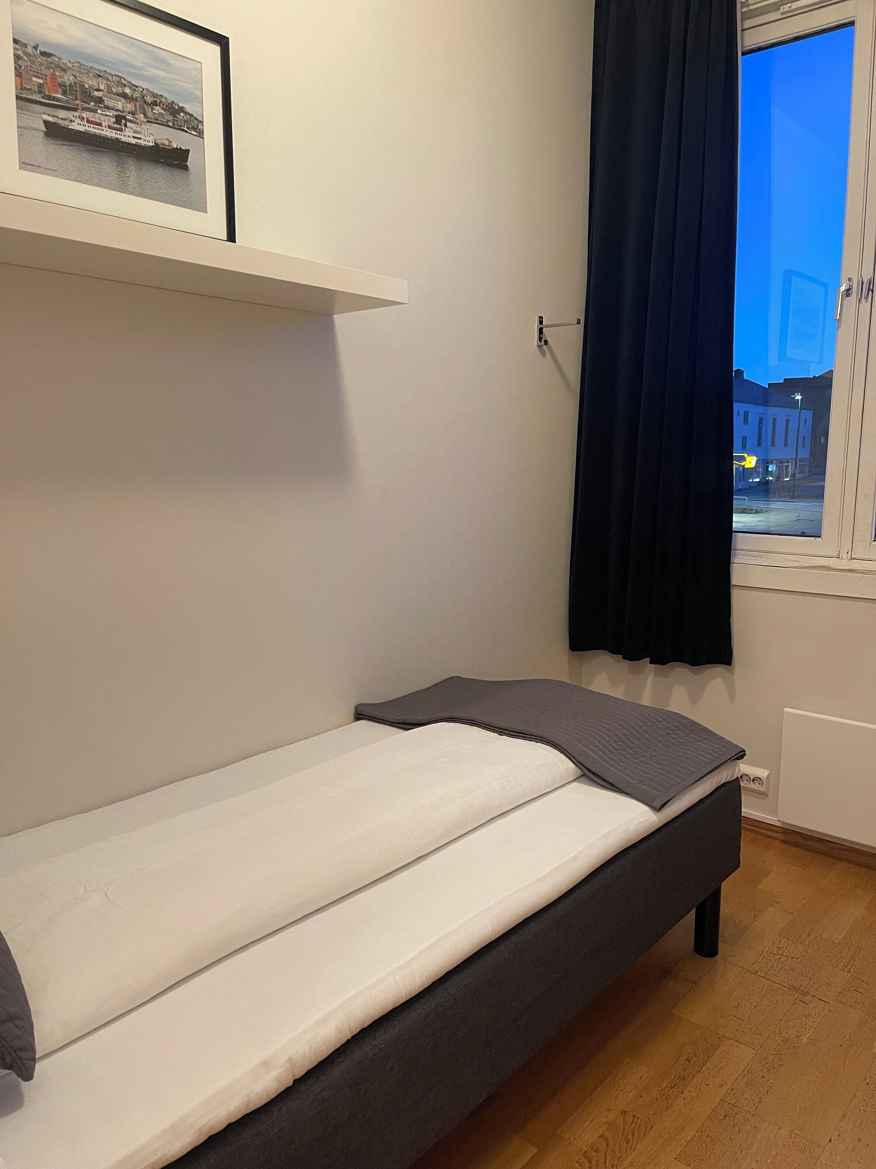 A small, single-bed room with a white mattress and dark headboard. A framed picture hangs on the wall above the bed.