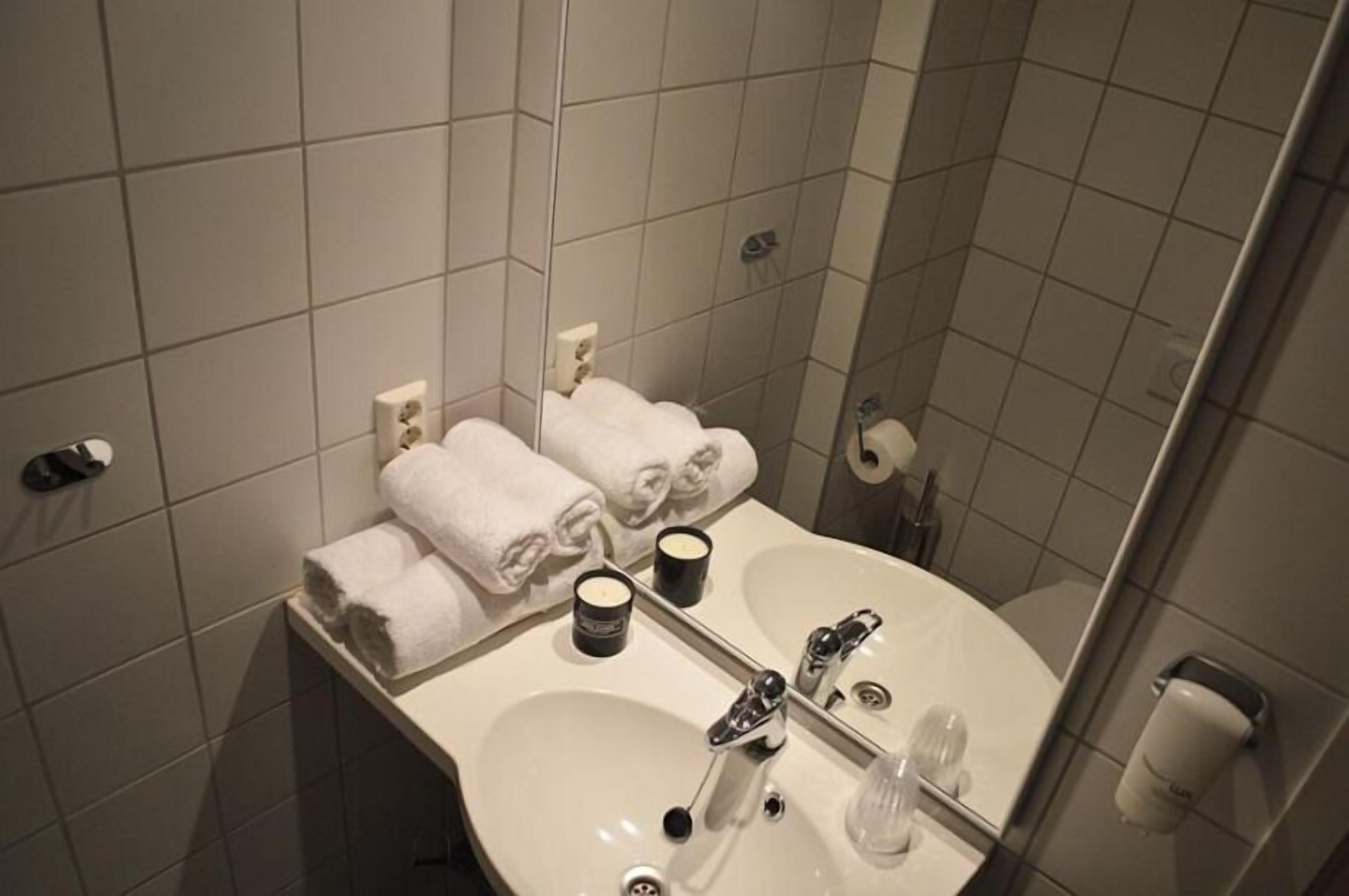 Casa de banho com lavatório branco, duas torneiras, espelho e porta-toalhas com toalhas dobradas.