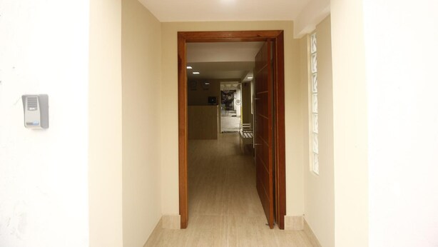 A hallway with a wooden door leading to a room. There is a light switch on the wall and a small device on the left. - Hotel Interlagos Xua (São Paulo)