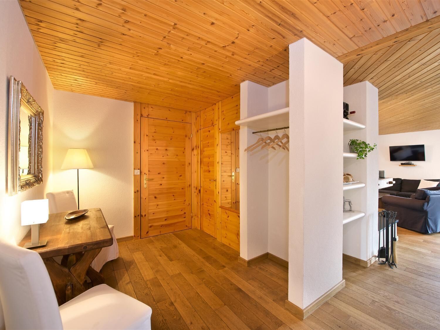 Ein gemütliches Zimmer mit Holzboden, Holzbalkendecke, einer weißen Wand, einem kleinen Tisch mit Lampe, einem Sessel, einem Fernseher und einem Sofa.