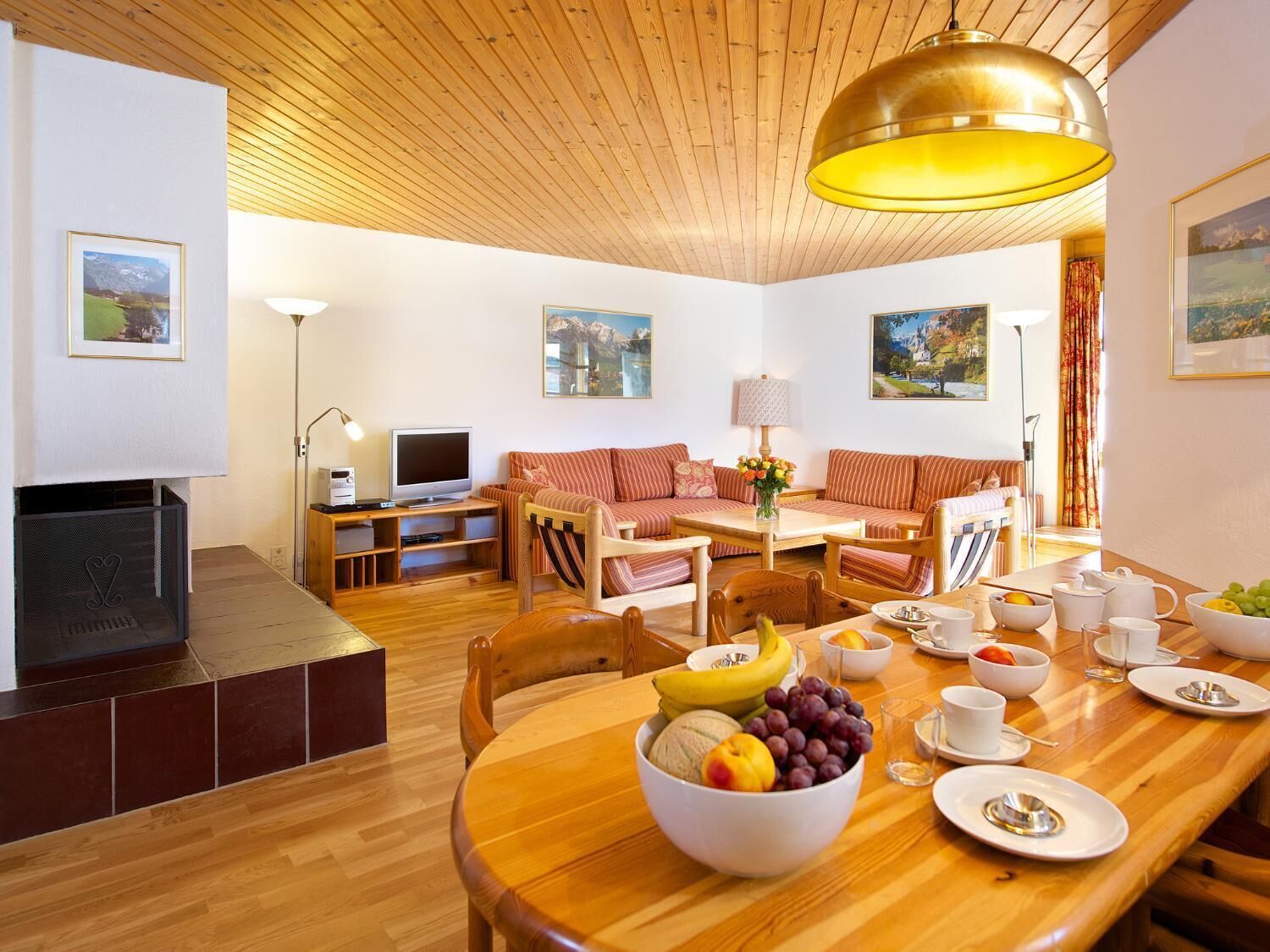 A cozy living room with a wooden ceiling, a television, a sofa, and a dining table set with fruits and cups.