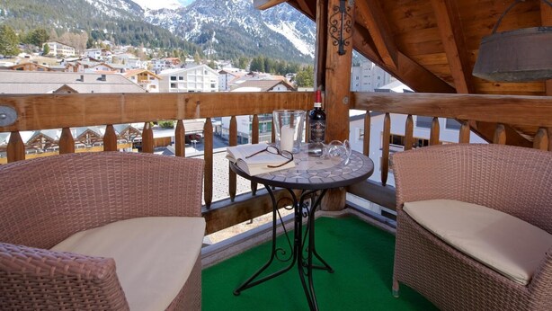 A balcony with wicker furniture, a table, and a view of a mountain town.