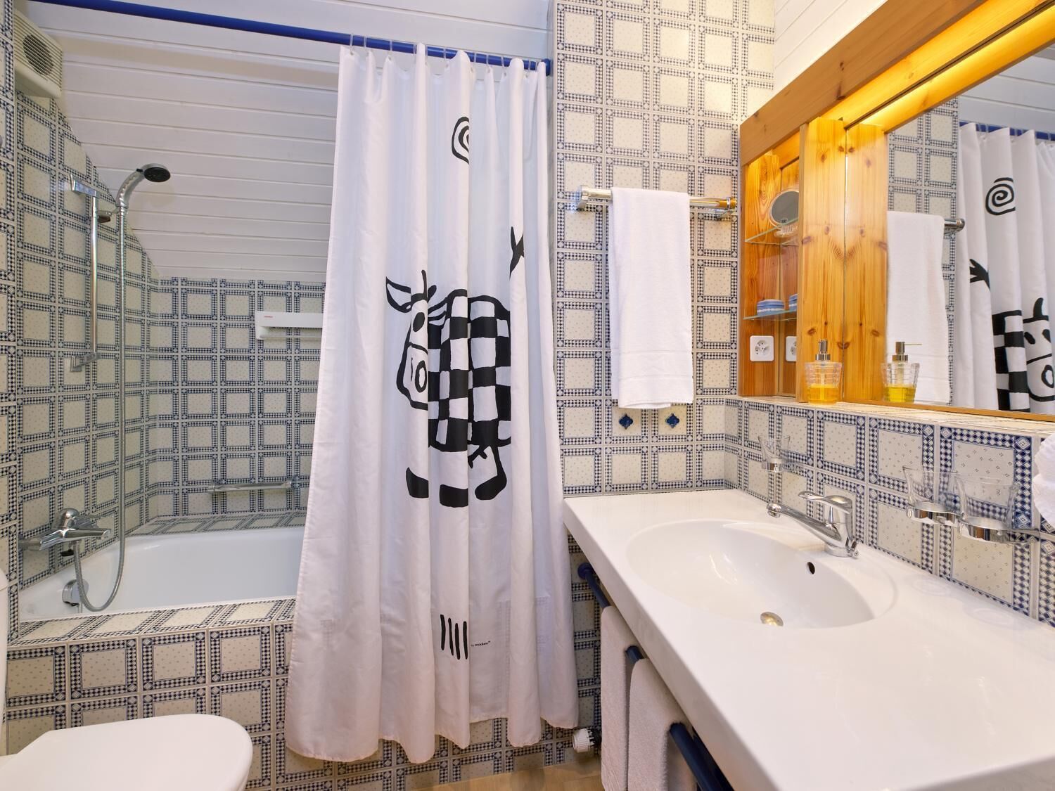 Ein Badezimmer mit fliesenbesetzter Rückwand, einem weißen Waschbecken, einem Spiegel mit Holzrahmen und einer Duschvorhang mit Kuhmuster.