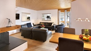 A modern living room with a wooden ceiling, a sofa set, a dining table with chairs, and a fruit bowl on the table.