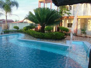 Una piscina con caída de agua, rodeada de follaje cuidado y un edificio con balcones.