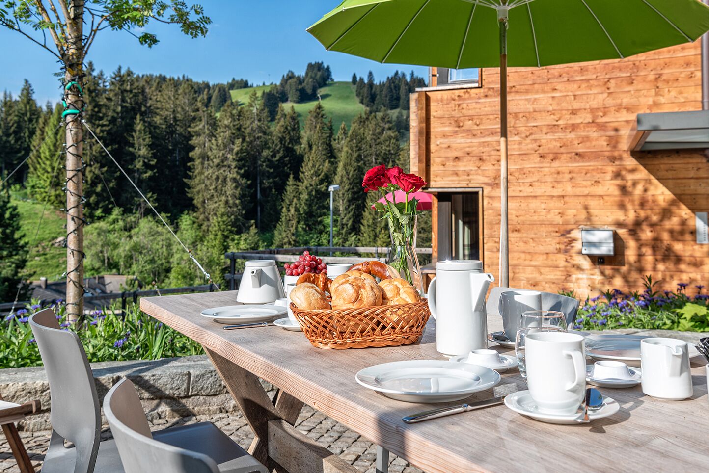 A wooden table set with breakfast items, a wicker basket, and a vase with red flowers, overlooking a forested area.