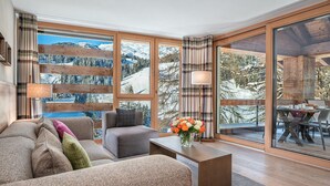 A cozy living room with a sofa, armchair, and coffee table, large windows with a snowy mountain view, and a dining area with a table and chairs. - Turrabuel (Churwalden)