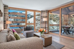 Un salón acogedor con sofá, sillón y mesa de centro, ventanas amplias con vistas a una montaña nevada, y un comedor con mesa y sillas.
