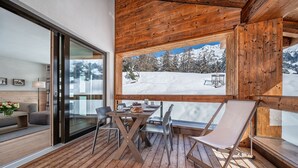 A wooden deck with a table and chairs, a sofa, and a large window overlooking a snowy mountain landscape.