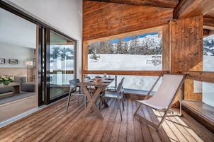 Una terraza de madera con una mesa, sillas, un sofá y un amplio ventanal con vistas a un paisaje montañoso nevado.