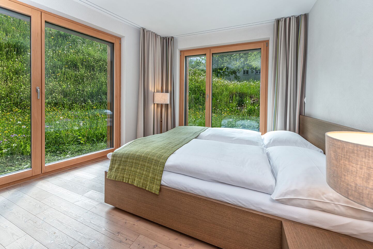 A bedroom with a large bed, a bedside lamp, and a view of greenery through floor-to-ceiling windows.