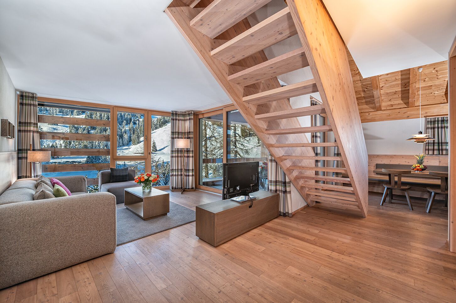 Un salon moderne avec un escalier en bois, de grandes fenêtres offrant une vue sur un paysage enneigé, un téléviseur à écran plat et un coin repas.