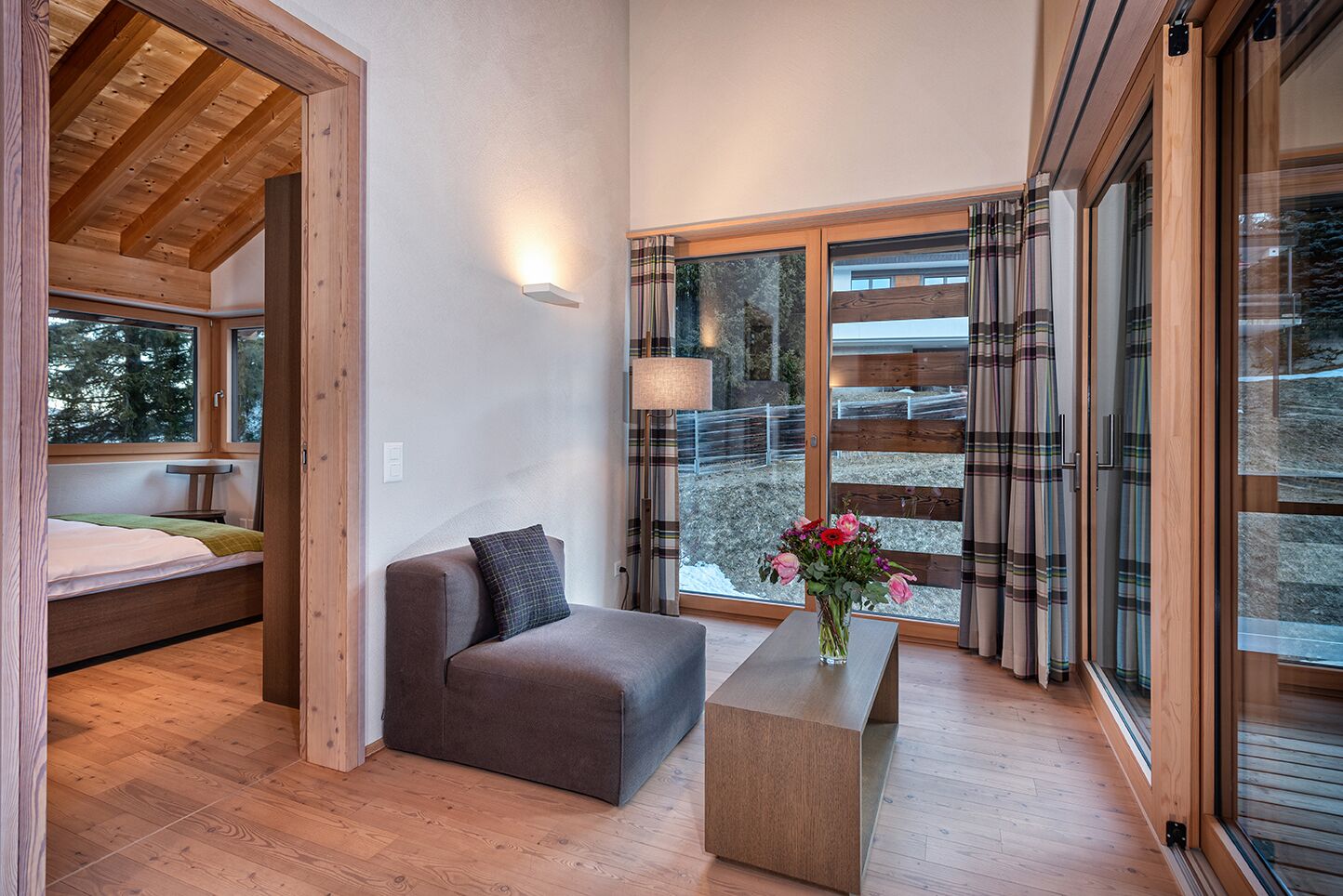 Un interior moderno con techo de madera, un sofá, una mesita con un florero y ventanas grandes con cortinas.