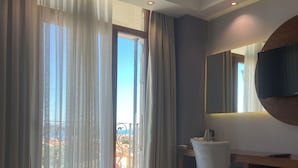 A hotel room with a large bed, a desk with a chair, a round mirror, and a view of the outdoors through sheer curtains. - Maison Vourla Hotel (Urla)