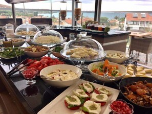 Un bufé con diversos platos, entre ellos ensaladas, quesos y carnes, dispuesto en una mesa con vista al mar.