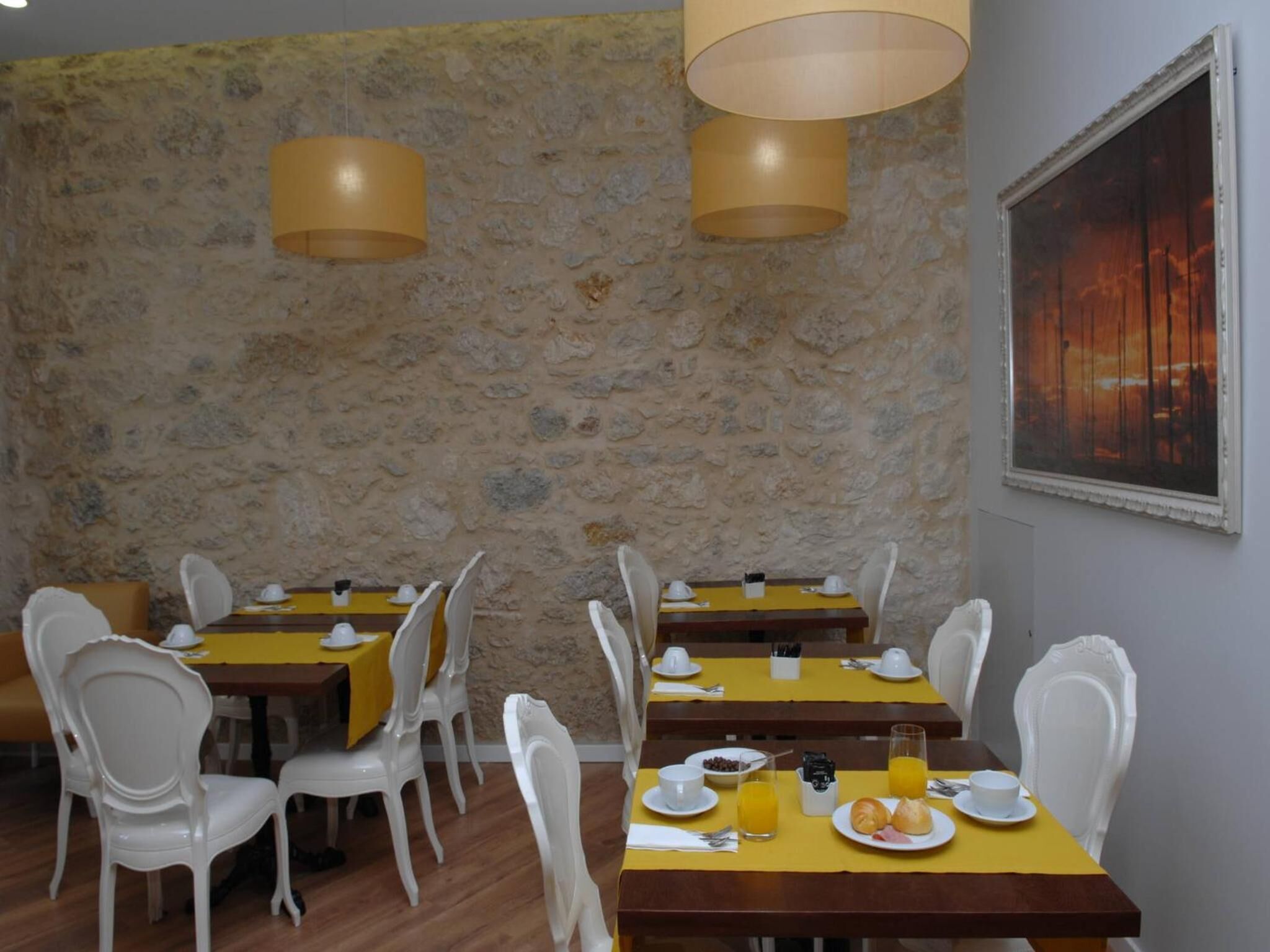 Comedor con paredes de piedra, sillas blancas, manteles amarillos y una pintura de atardecer.