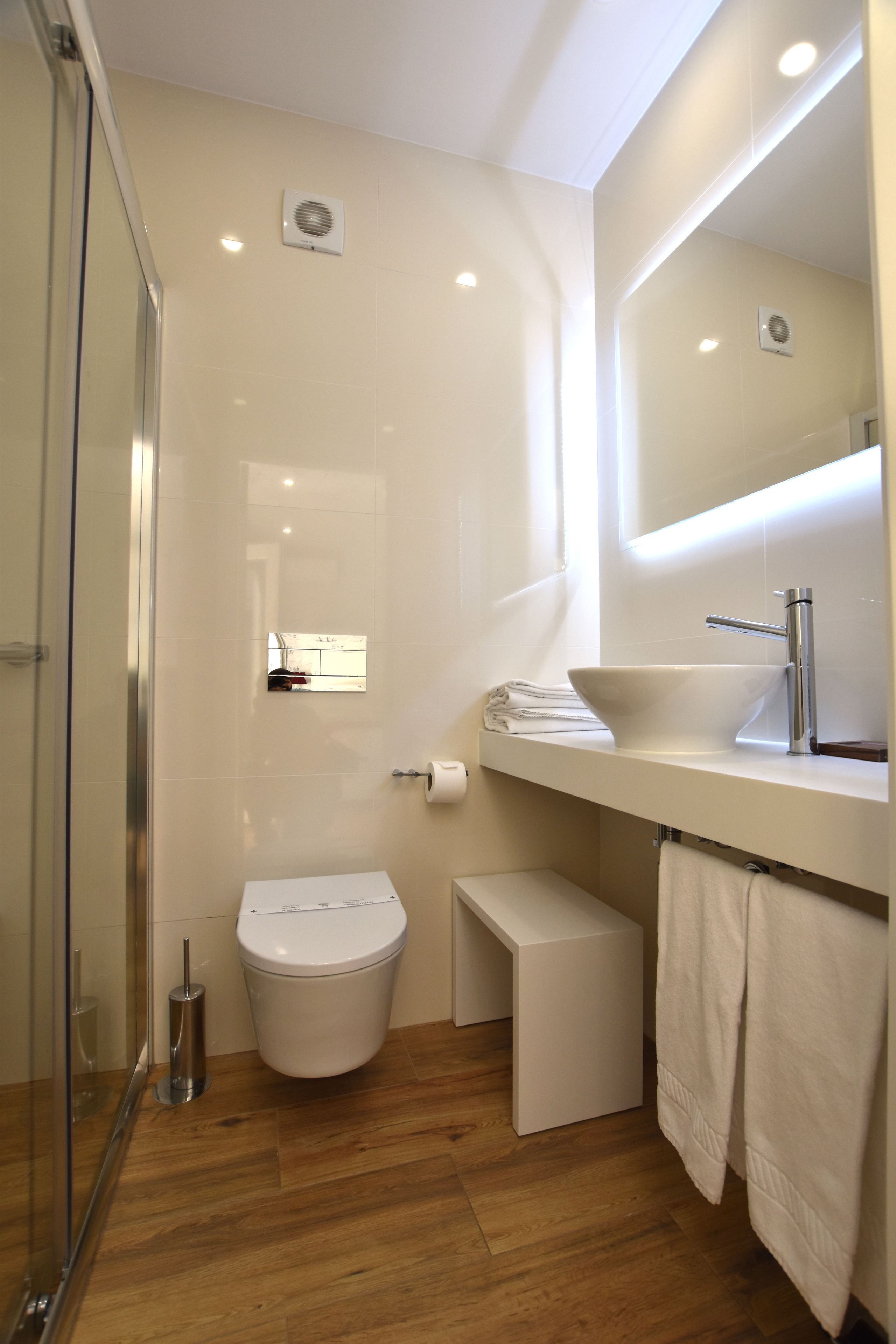 A modern bathroom with a glass shower enclosure, a white toilet, a sink with a mirror, and a small bench.