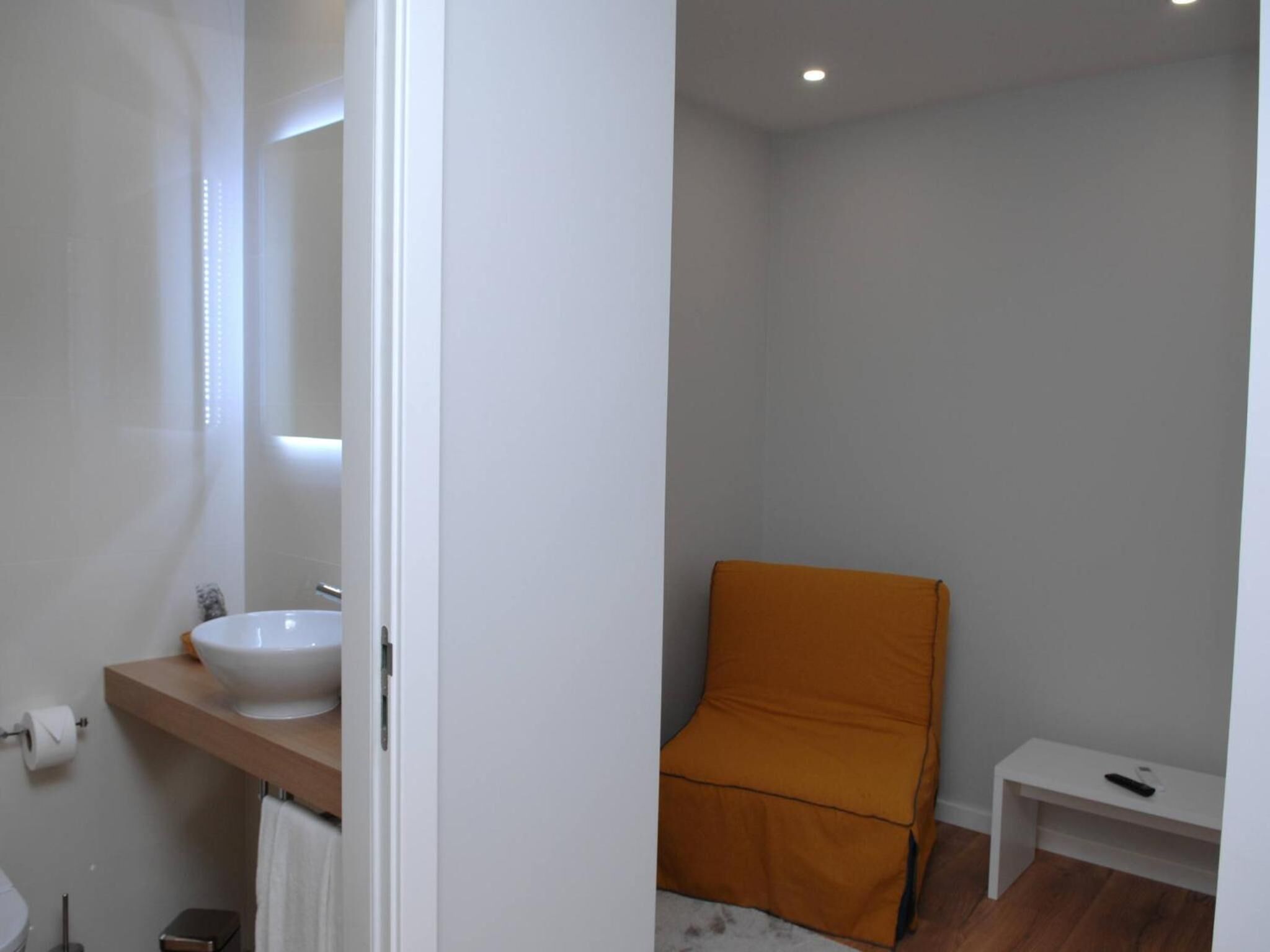 A bathroom with a white sink, a mirror, and a wooden shelf. There is a white toilet and a small white shelf with a remote control.