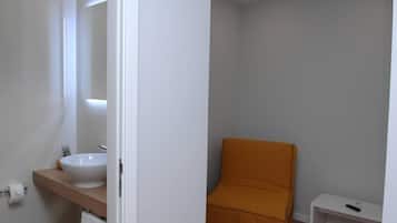 Un baño con un lavamanos blanco, un espejo y un estante de madera. Hay un inodoro blanco y un pequeño estante blanco con un control remoto.