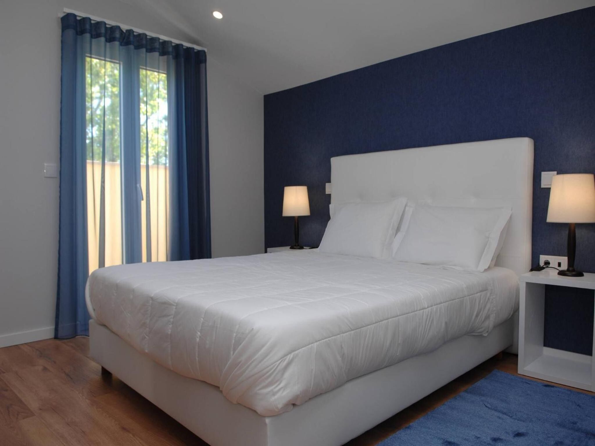 Un dormitorio con una cama blanca, dos mesitas de noche con lámparas, una pared azul y suelo de madera.
