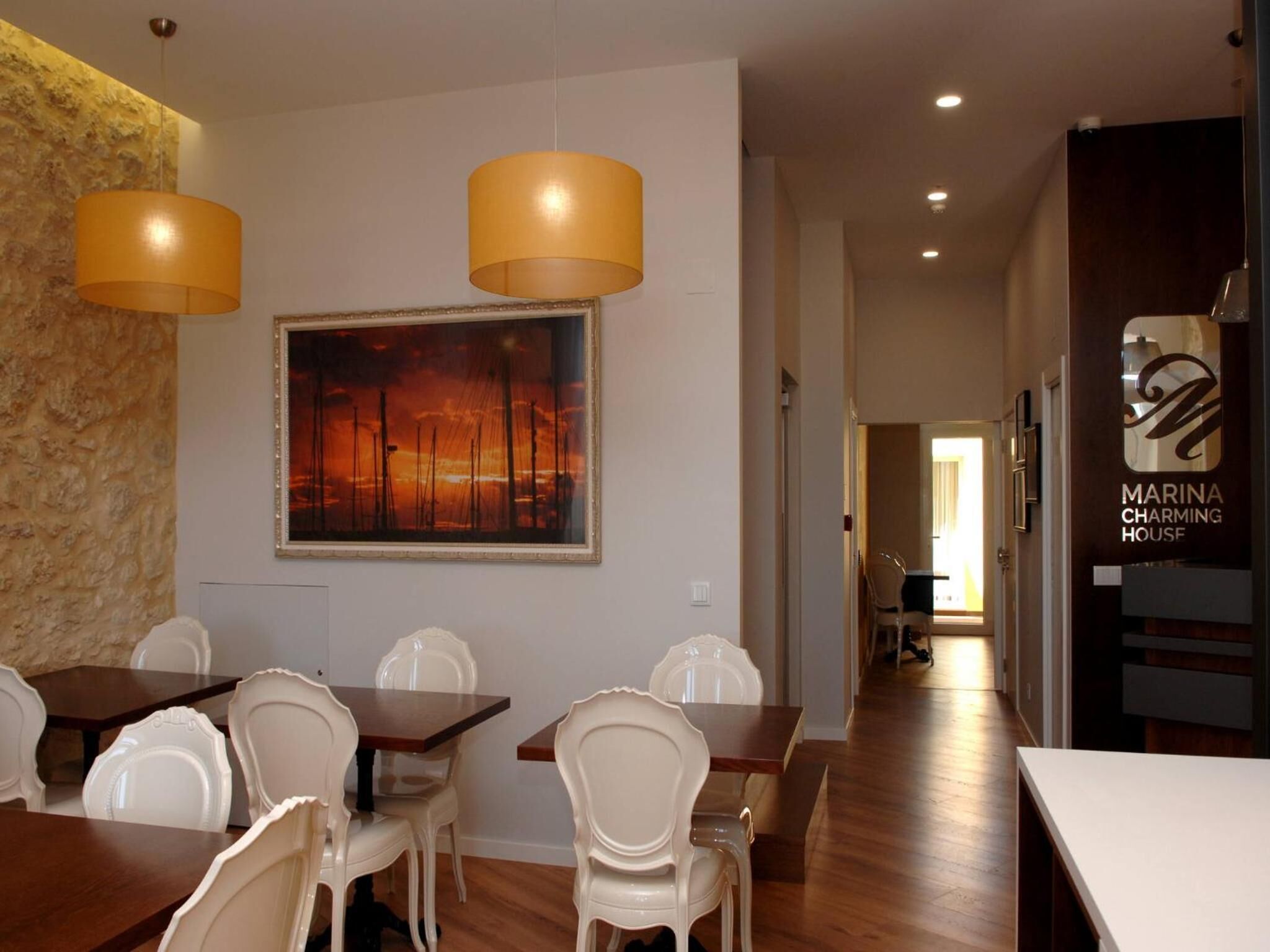 A dining area with white chairs and wooden tables, a large painting of a sunset, and a sign for 'Marina Charming House'.