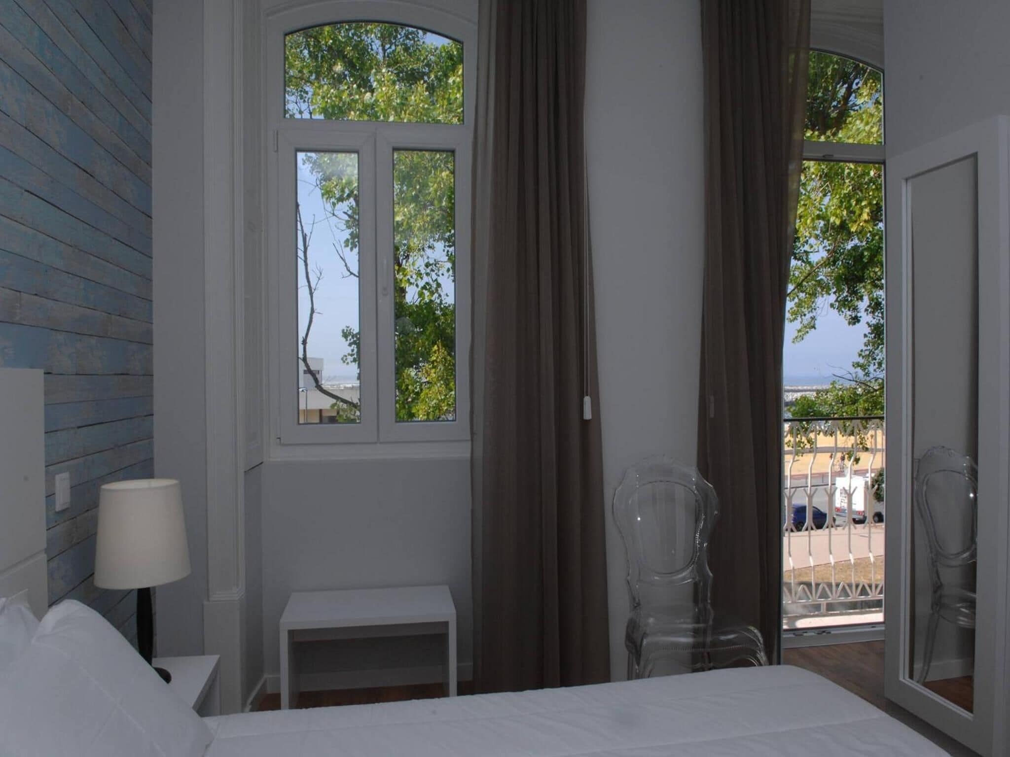 A bedroom with a bed, a small bedside table, a lamp, and a chair. There are two windows with curtains, one of which has a view of trees and a building.