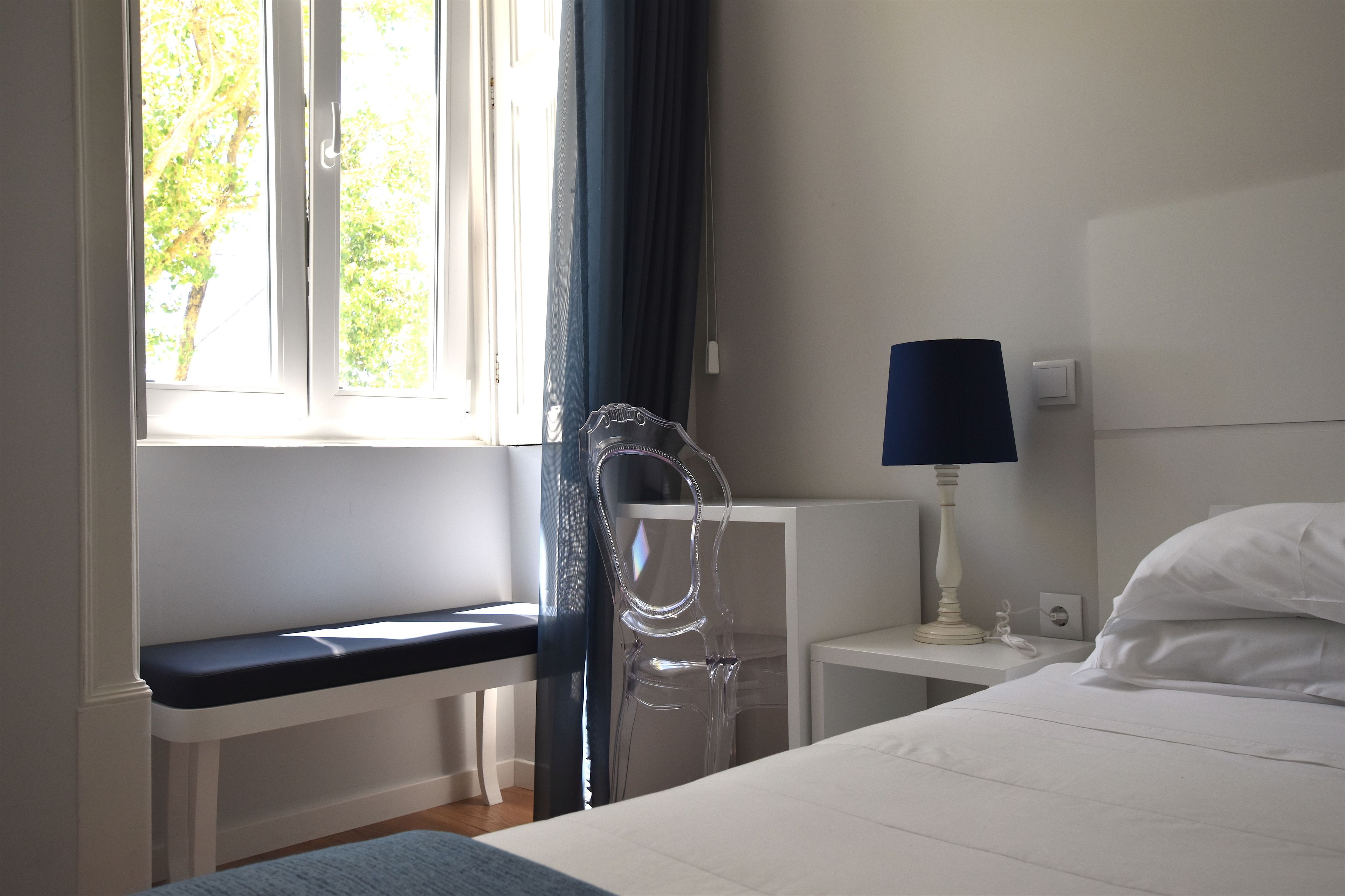 A bedroom with a bed, a bedside table with a lamp, a chair, a window with curtains, and a bench by the window.