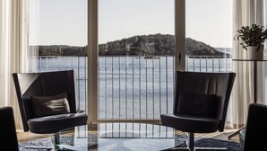 A room with a large window overlooking a body of water and a distant hill. There are two black chairs with cushions, a small table, and a potted plant on a side table. - Destination Bokenäset (Uddevalla)