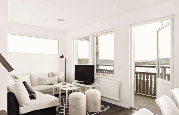 A modern living room with a sofa, ottomans, a TV, and large windows offering a view of a body of water.