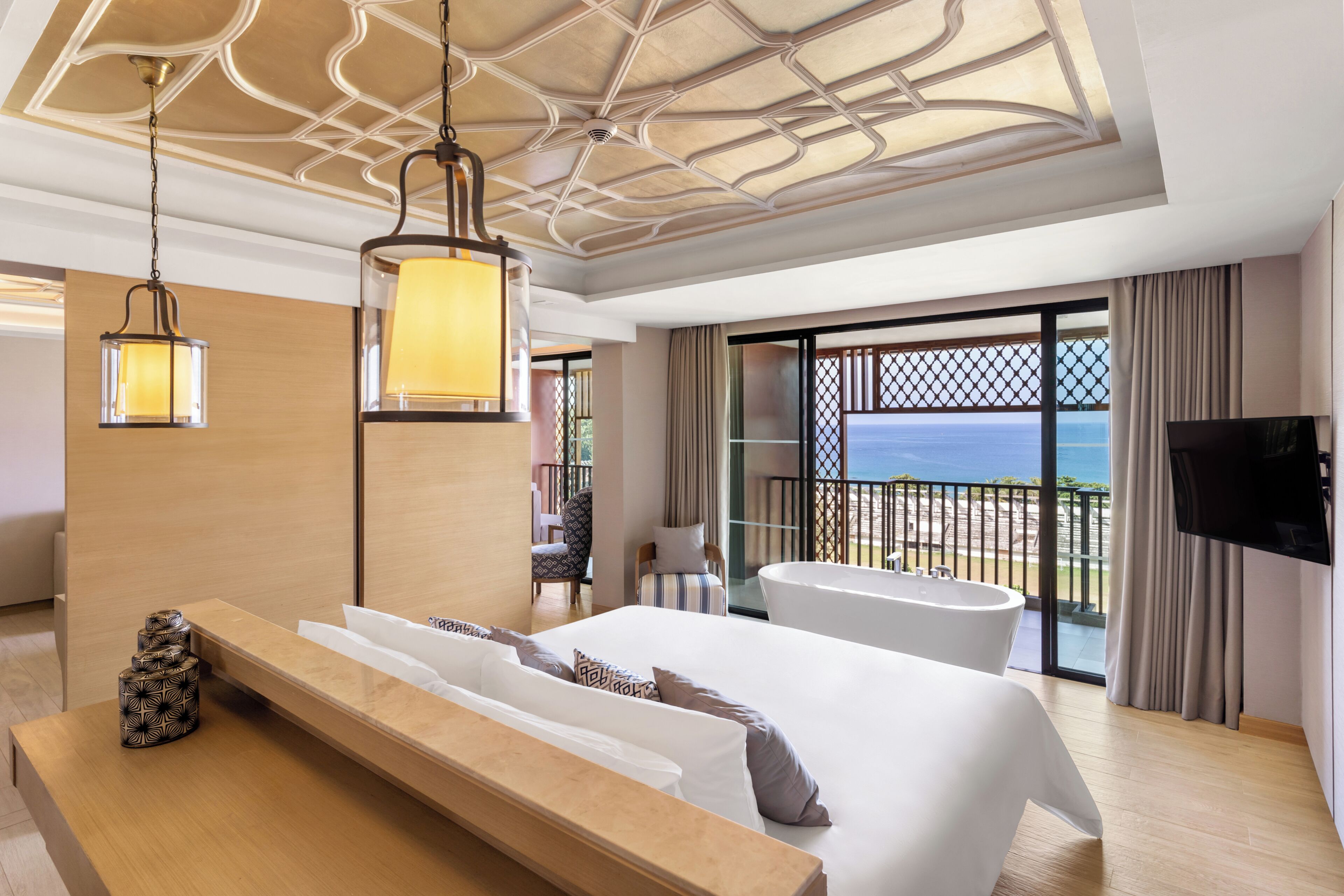 A modern hotel room with a large bed, a sitting area, a bathtub, and a balcony with a view of the sea.