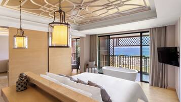A modern hotel room with a large bed, a sitting area, a bathtub, and a balcony with a view of the sea.