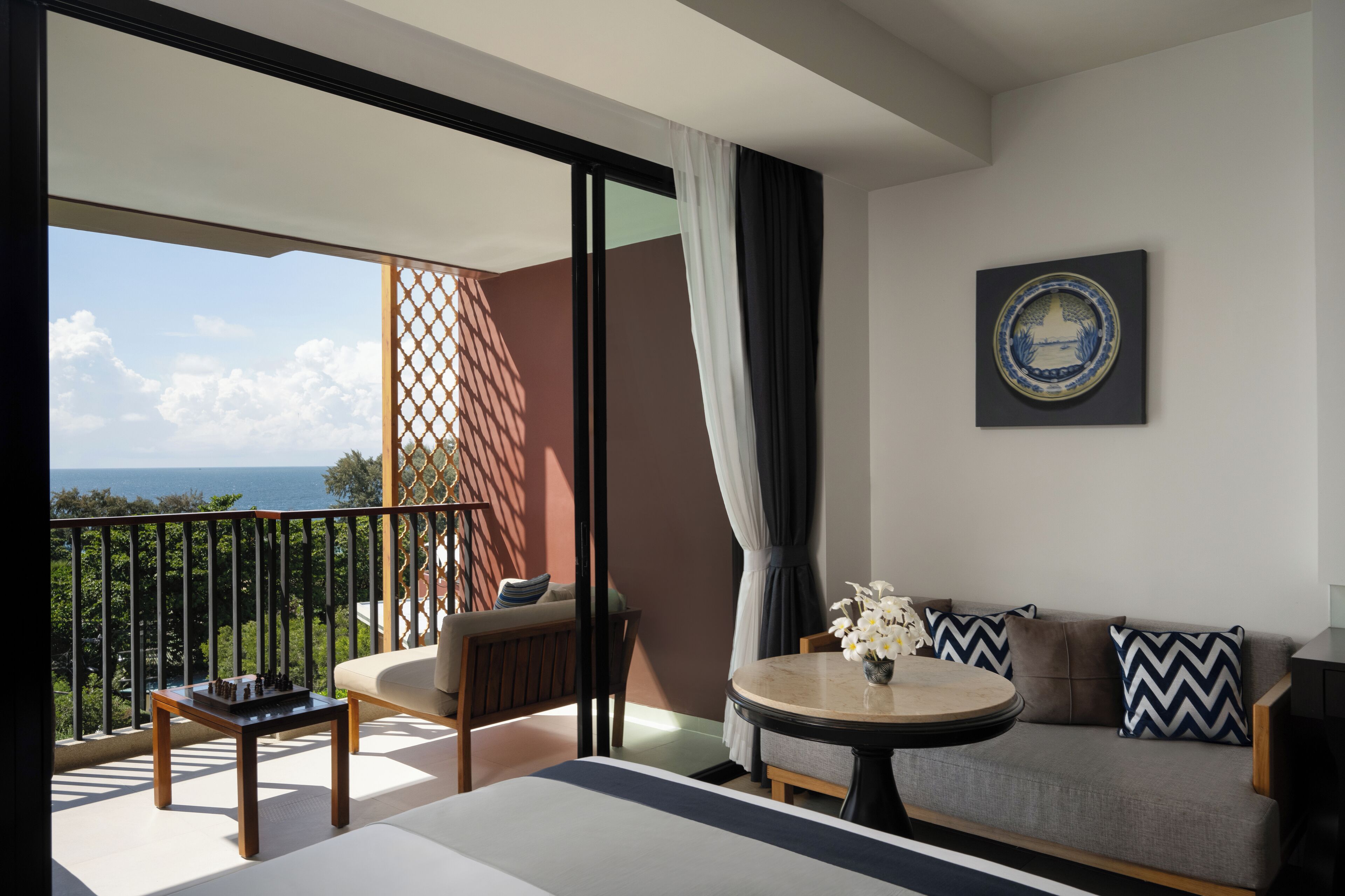 Een balkon met uitzicht op zee, een bank, een tafel met een bloevas en een decoratief bord aan de muur.
