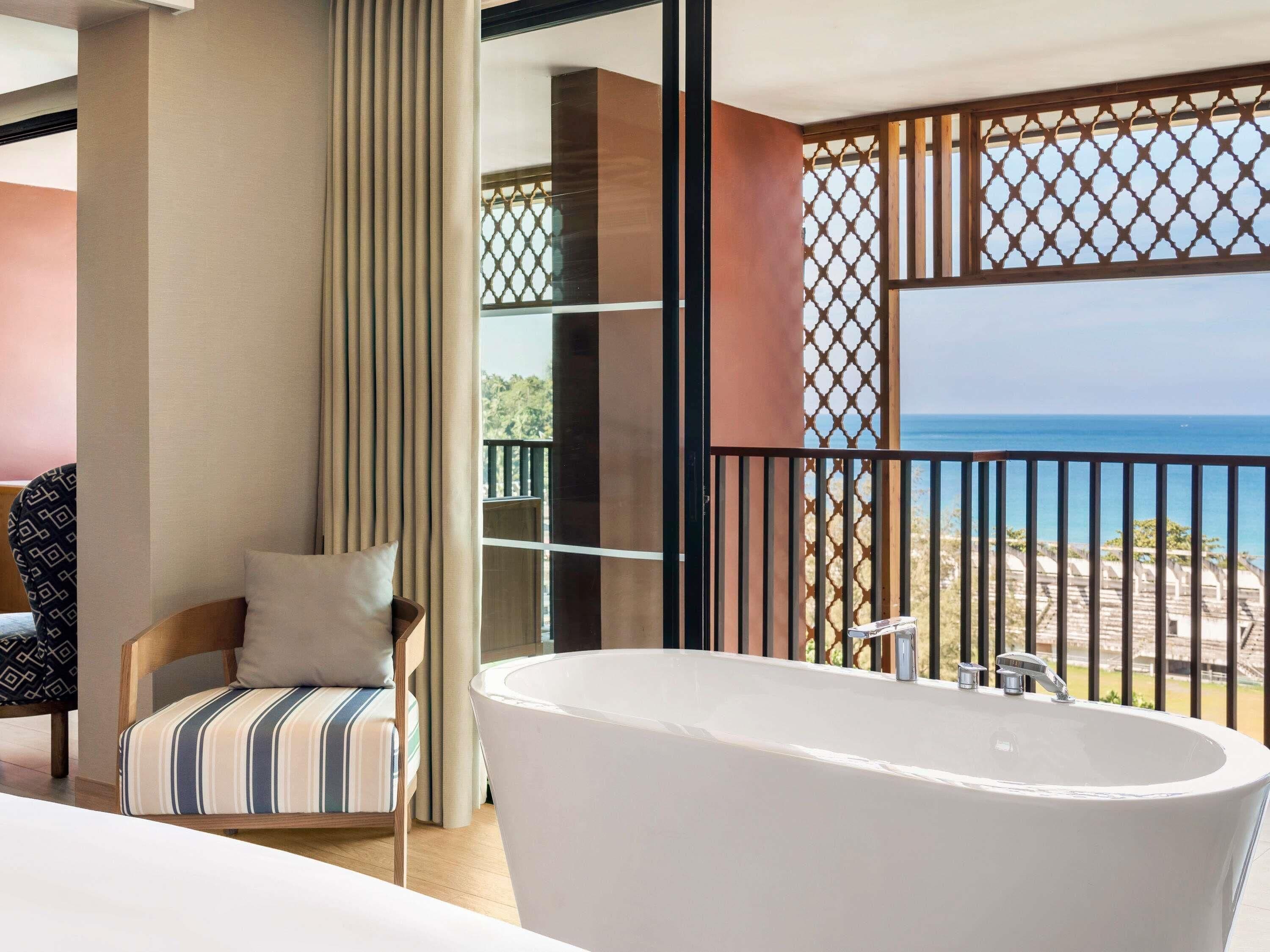 A modern bathroom with a freestanding bathtub, a chair with a striped cushion, and a view of the sea through a balcony with a lattice design.