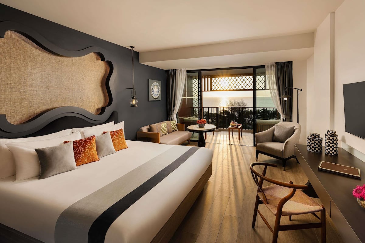 A hotel room with a large bed, a sofa, a dining table, and a balcony with a view.