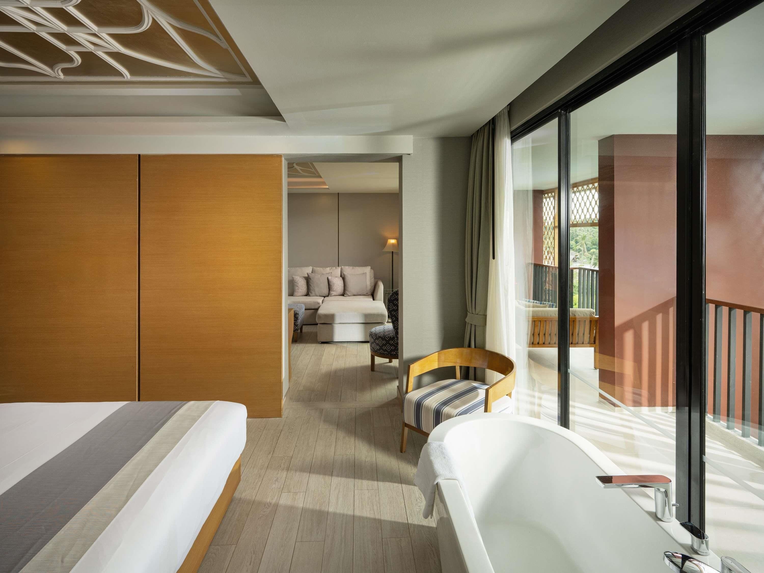 A modern hotel room with a large bed, a separate seating area with a sofa and chairs, a balcony with a railing, and a large window.