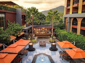A rooftop bar with tables and chairs, surrounded by greenery and a view of hills.