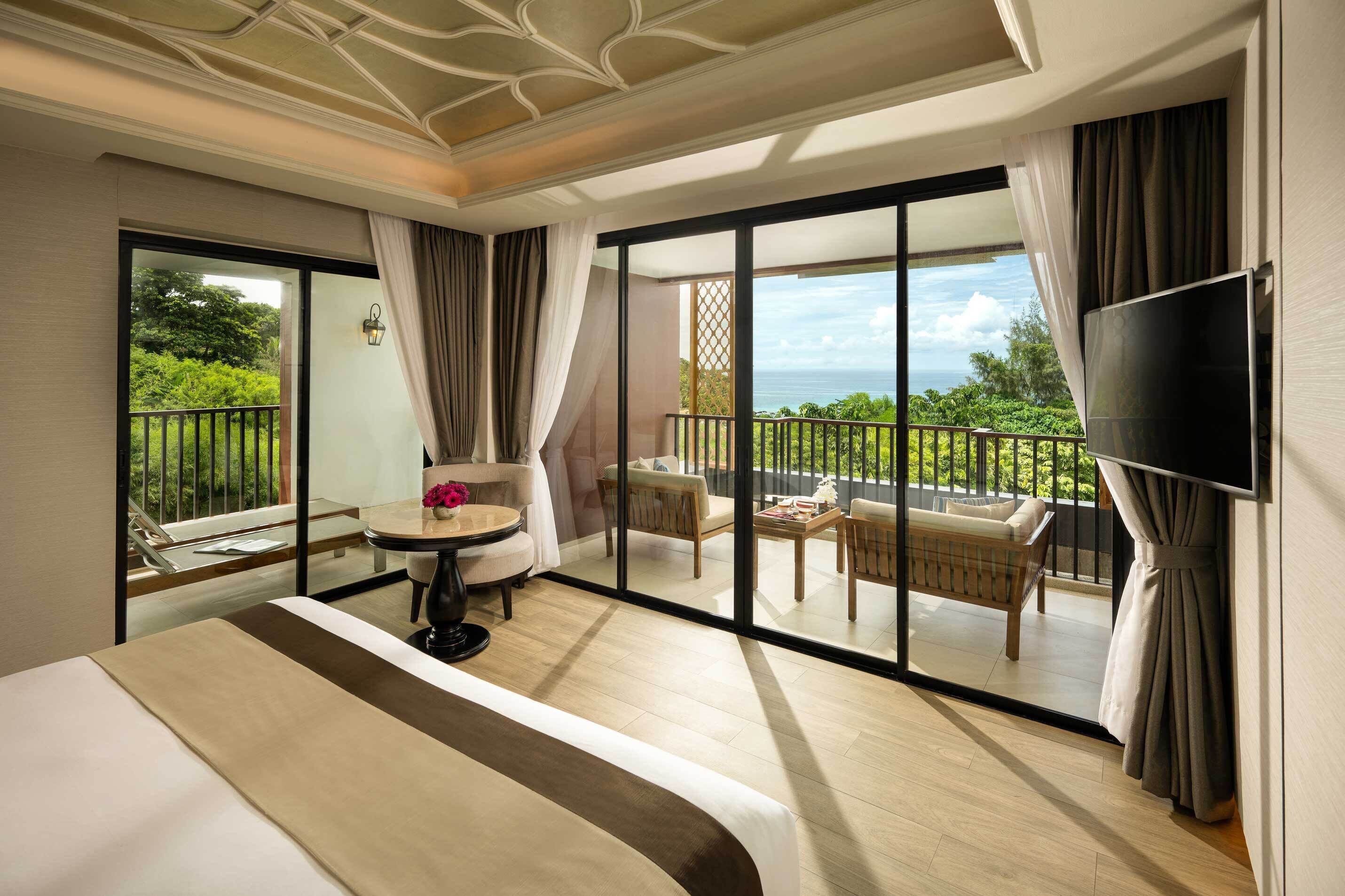 A hotel room with a large bed, a small table with a vase, a sofa, and a balcony with a view of the sea.