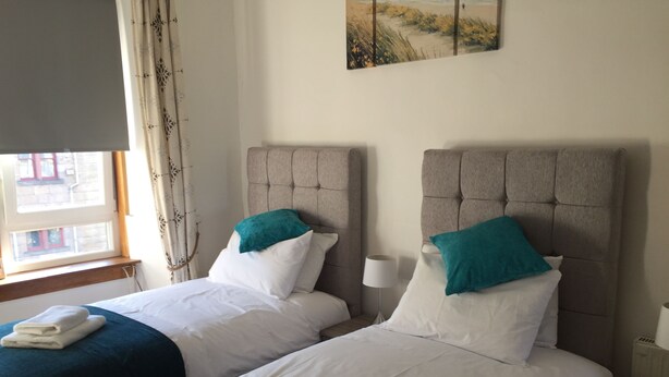 A hotel room with two beds, each with a headboard, white bedding, and teal pillows. There is a window with curtains, a bedside lamp, and a painting on the wall.