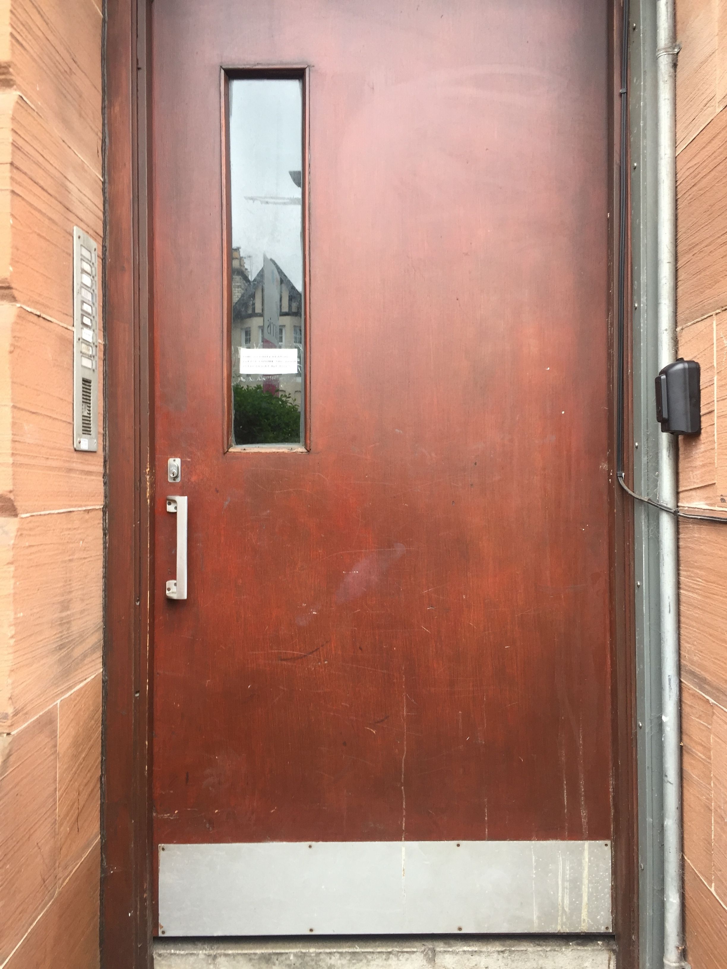 A wooden door with a small rectangular window, a metal handle, and a metal plate on the left side.