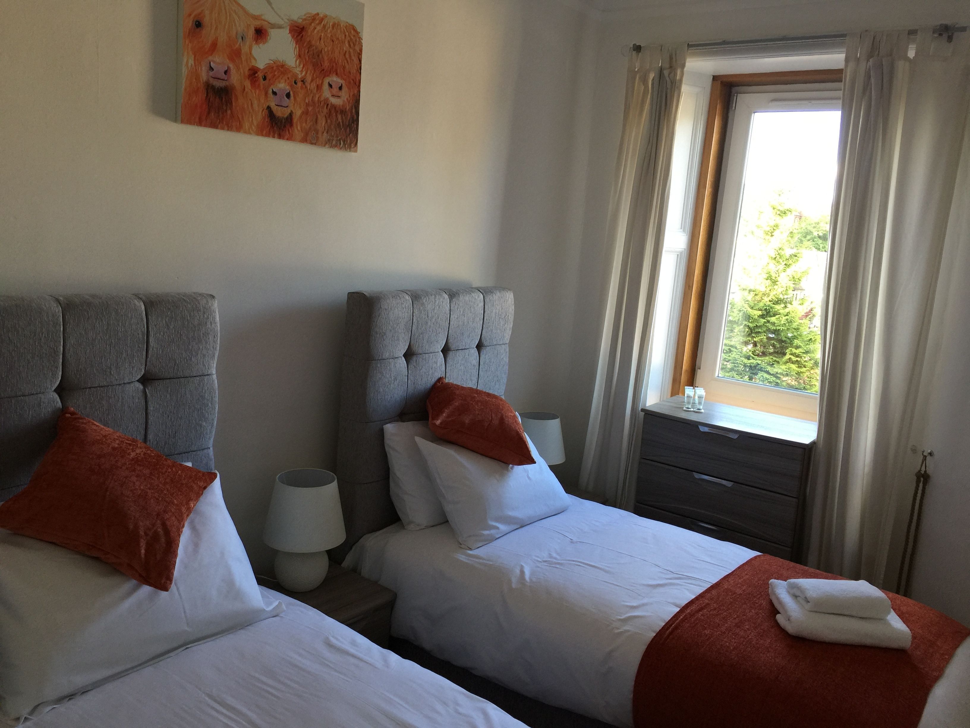 A hotel room with two beds, a headboard, a nightstand, a lamp, and a window with curtains.