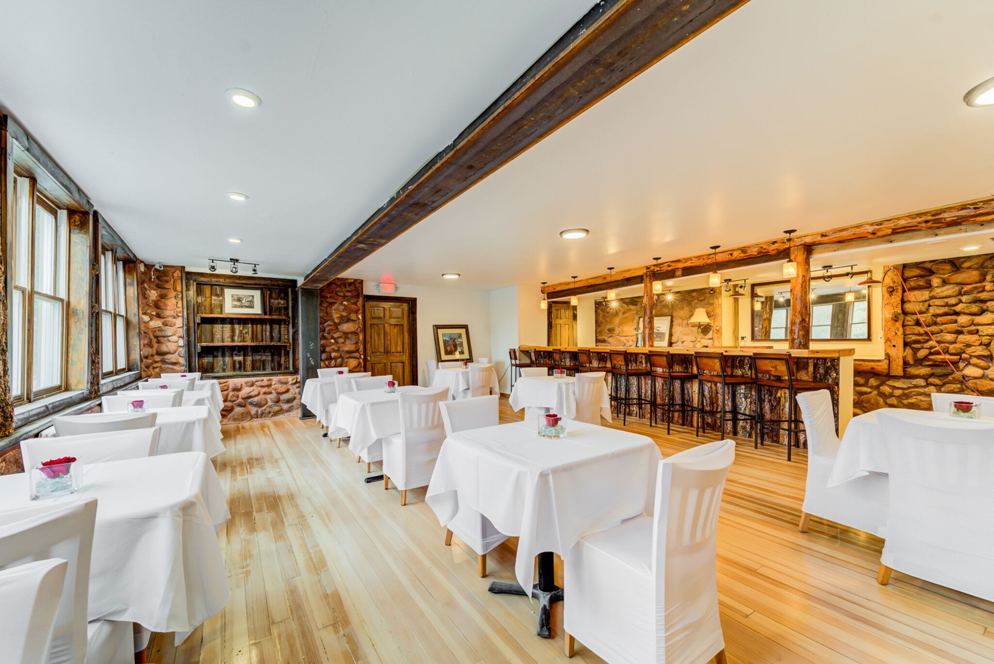 A restaurant with multiple tables and chairs, stone walls, wooden beams, and a bar area.