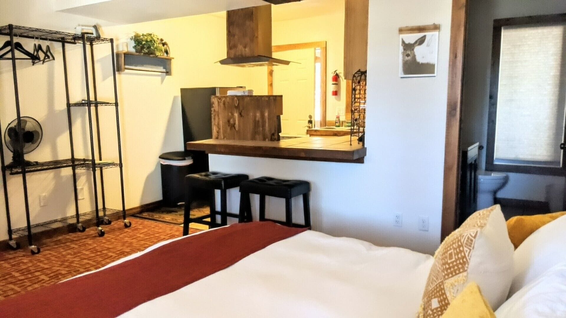 Une chambre d’hôtel avec un lit, un bureau avec deux chaises, un miroir, une étagère avec des plantes et une fenêtre donnant sur un paysage.