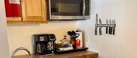 A compact kitchen with wooden cabinets, a stainless steel sink, a microwave, a coffee maker, and a set of knives on the wall.