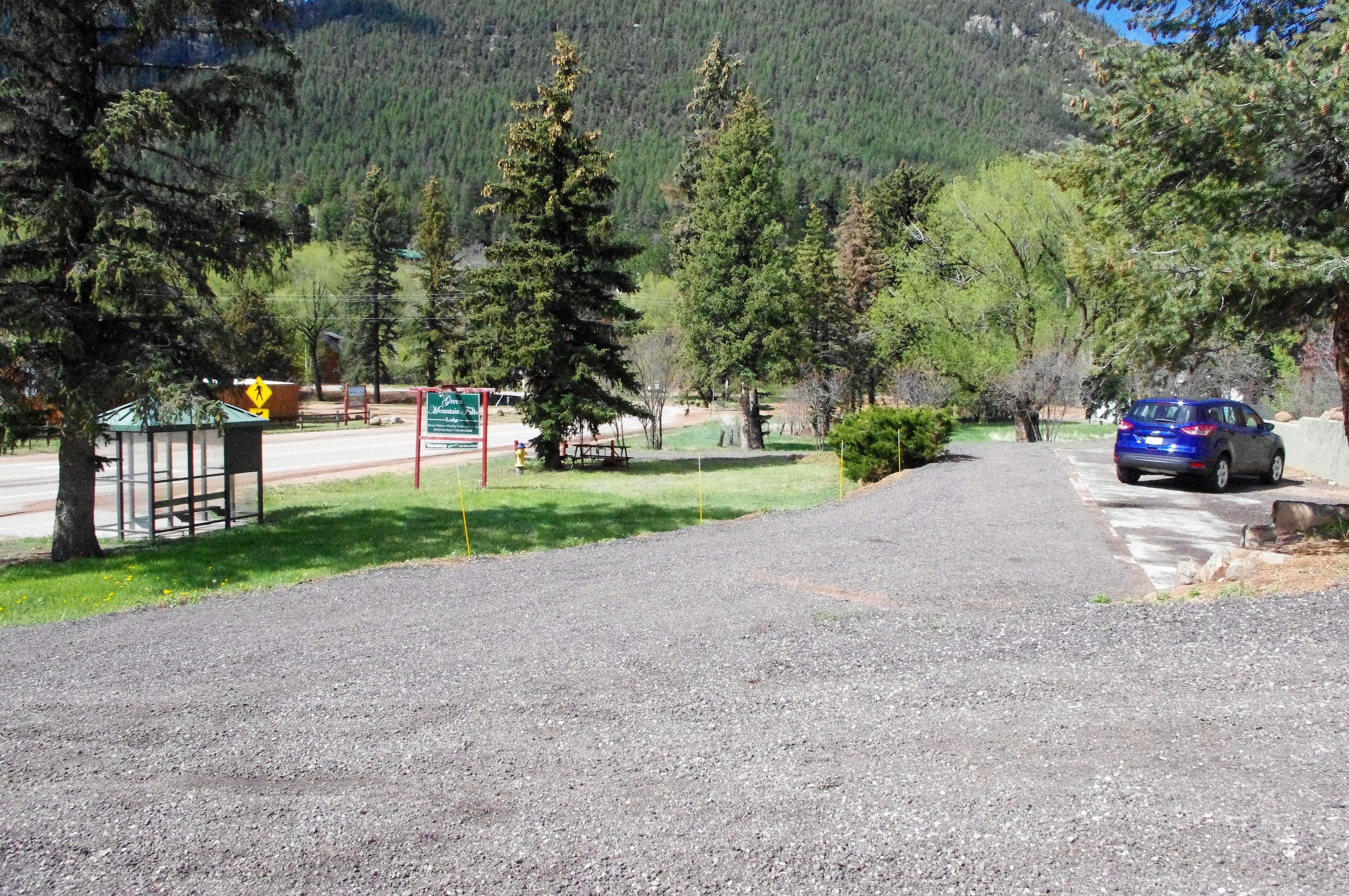 A gravel road leading to a parking area with a blue car parked on the right. There are trees and a mountain in the background.