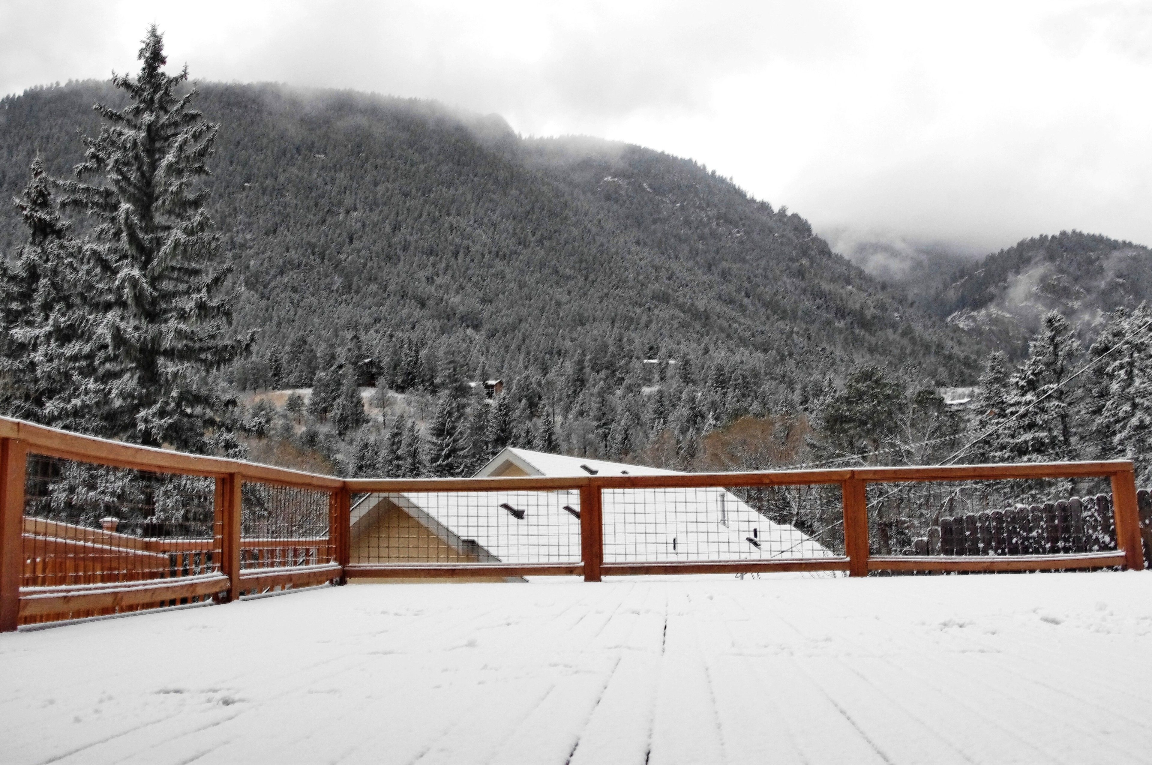 Une terrasse enneigée, dotée d’une rambarde en bois, qui offre une vue sur une région montagneuse couverte de forêts.