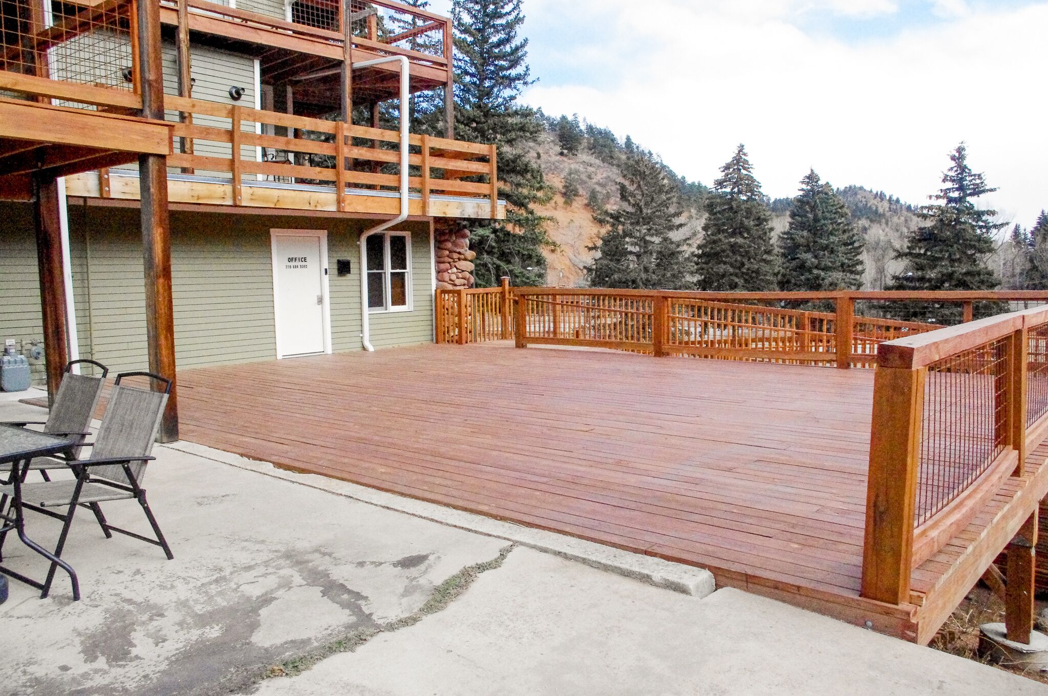 Une terrasse en bois avec une rambarde, une table avec des chaises et un bâtiment dont la porte est marquée du mot « BUREAU ».
